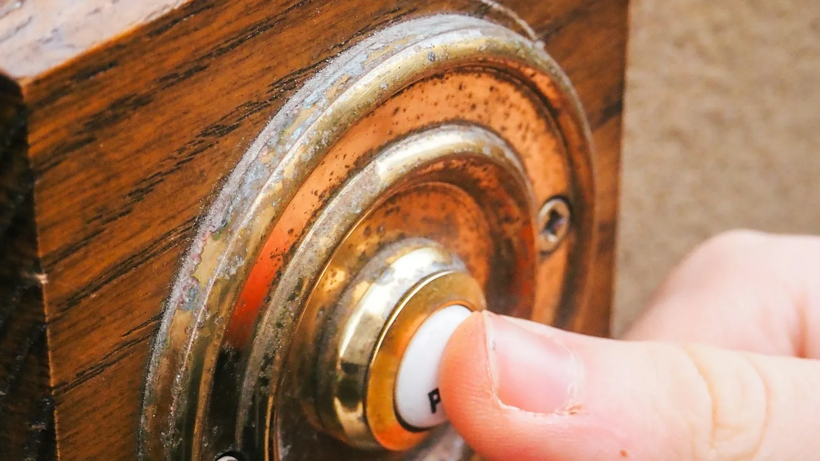 A finger pressing a brass doorbell button on a wooden door frame